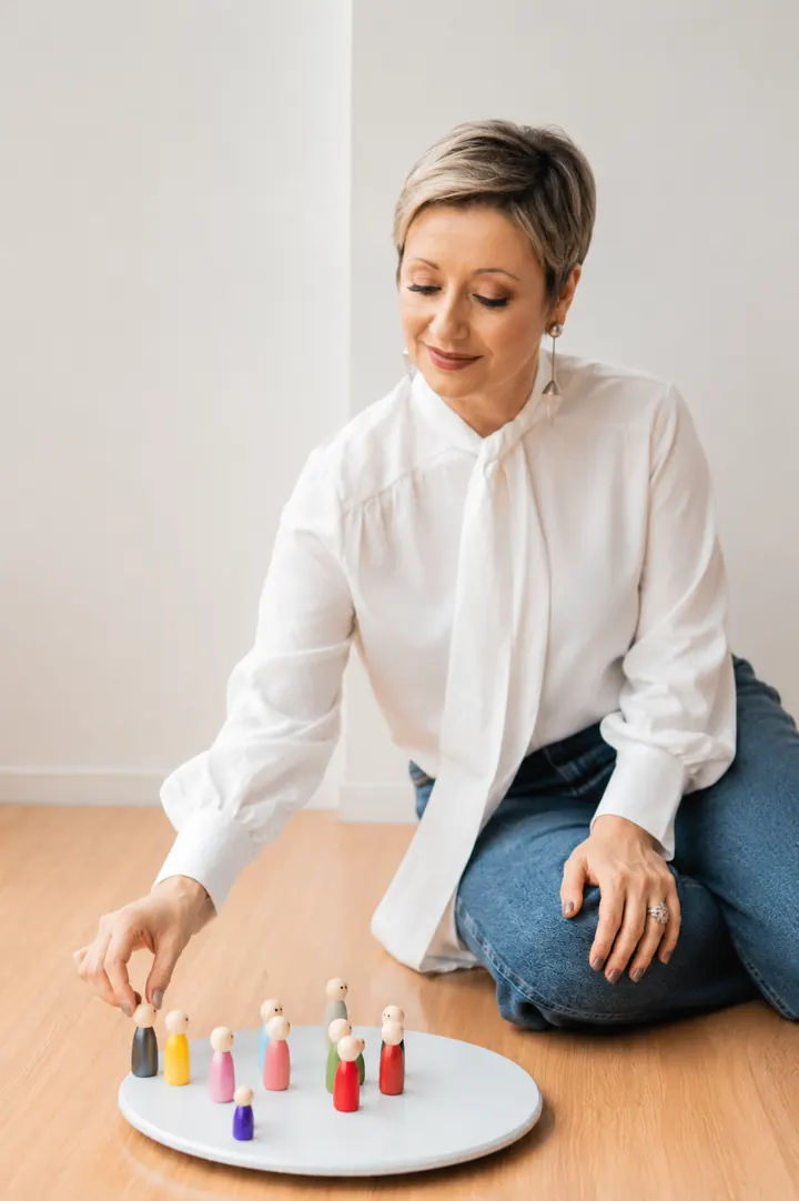 Tatti Santos sentada no chão, posicionando bonecos coloridos de constelação sobre uma mesa redonda.