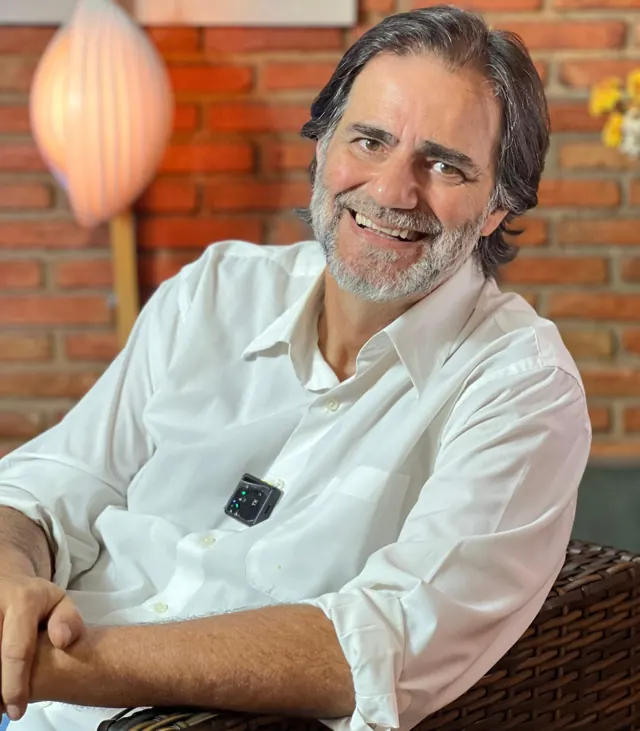 Tadeu Ohl sorrindo, de camisa branca, sentado em frente a uma parede de tijolos.